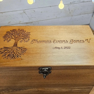 May include: A rectangular wooden box with a hinged lid and a metal latch. The lid features a laser-engraved tree with visible roots on the left side, and the text "Thomas Evans Jones V" followed by the date "May 5, 2022" on the right side.