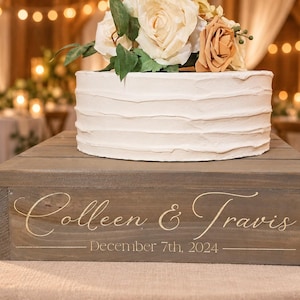 May include: A square wooden cake stand with a grey finish, featuring the white printed text Colleen & Travis and December 7th, 2024. A round white frosted cake topped with cream and peach-colored artificial flowers sits on the stand.