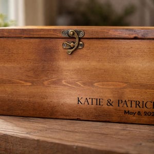 May include: A rectangular wooden box with a hinged lid and a metal clasp. The box is stained a warm brown color and features the engraved names "KATIE & PATRICK" and the date "May 8, 2026".