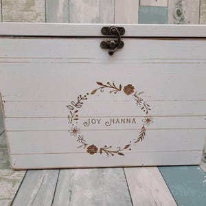 May include: A rectangular white wooden box with a hinged lid and a bronze-colored metal latch. The front features a laser-engraved floral wreath design surrounding the name JOY HANNA in a simple, clean font. The wood has a distressed finish.
