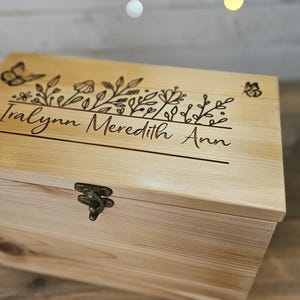 May include: A rectangular light-colored wooden box with a hinged lid and a metal latch. The top of the lid features a laser-engraved design of wildflowers and butterflies, with the name Iralynn Meredith Ann engraved in a script font.