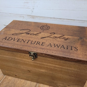 May include: A rectangular brown wooden box with a hinged lid and a brass-colored metal latch. The lid features an engraved compass rose, the name Jack Lakes in script, and the text ADVENTURE AWAITS in a bold, capitalized font.