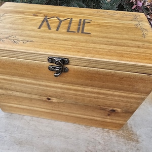 May include: A rectangular wooden storage box with a hinged lid and a dark metal latch. The top surface features the engraved name KYLIE in capital letters, flanked by delicate floral line art engravings on the left and right corners.
