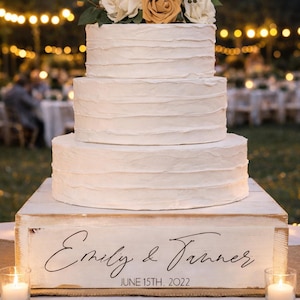May include: A three-tiered white wedding cake with textured frosting, topped with cream and gold roses and greenery. The cake sits on a white wooden box with the names "Emily & Tanner" and the date "June 15th, 2022" written on it. Lit candles flank the cake.