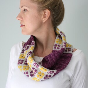 May include: A knitted infinity scarf with a geometric pattern. The scarf features a color block design with a deep purple base, a section with yellow and purple floral-like shapes on a light gray background, and a light gray section.