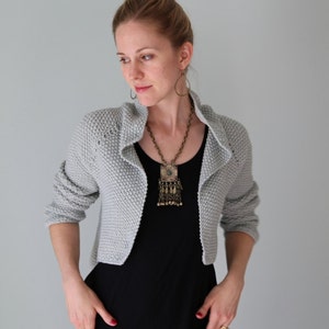 May include: A woman wearing a gray cropped cardigan with a black top and a long pendant necklace. The necklace has a large, ornate pendant with a tassel.