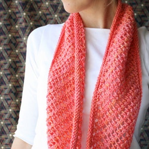 May include: A coral-colored knitted infinity scarf with a delicate openwork pattern. The scarf is made of soft yarn and has a subtle ombre effect.