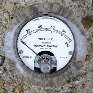 May include: A white analog voltmeter with a black needle, labeled "VOLTS D.C." and "Western Electric". The meter has a clear plastic cover and a black metal rim. The meter reads up to 100 volts and has a 1000 ohms per volt sensitivity.