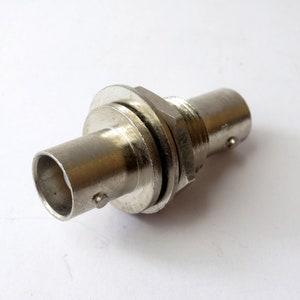 May include: A silver-colored BNC connector with a threaded nut and a female connector on one end and a male connector on the other end.