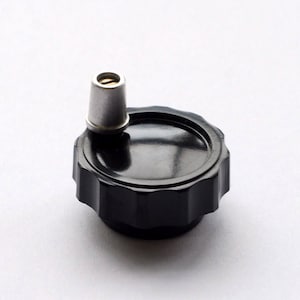 May include: A black plastic knob with a silver metal insert. The knob has a circular shape with a textured surface and a raised edge.