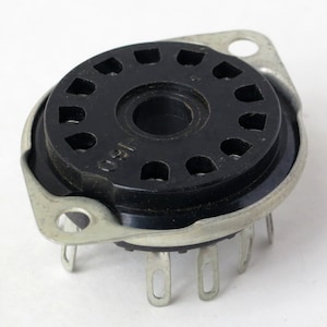 May include: A black and silver vintage vacuum tube socket with 10 pins. The socket has a circular base with a central hole and a ring of 10 holes around the edge. The socket is surrounded by a silver metal ring.