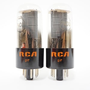 May include: Two vintage RCA vacuum tubes. Each tube has a clear glass enclosure with internal components. The base is black with orange RCA lettering and the letters "DT". The tubes are for audio equipment.