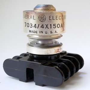 May include: A silver General Electric 7034/4X150A fuse with a black base and five terminals. The fuse is made in the U.S.A.