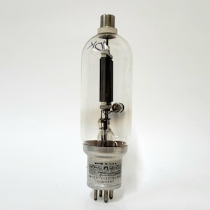 May include: A clear glass vacuum tube with a metallic base featuring the text MADE IN U.S.A. and UNITED ELECTRONICS COMPANY. The internal structure includes a dark rectangular component and a coiled wire element inside the transparent glass bulb.