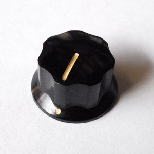 May include: A black, vintage-style guitar knob with a scalloped edge and a light-colored indicator line. The knob has a circular base and a small, light-colored dot on the side. This component is likely for a musical instrument.