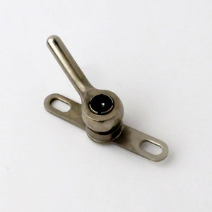 May include: A silver-toned metal window latch with a black plastic knob. The latch has a long arm that extends out to the side and a flat base with two holes.