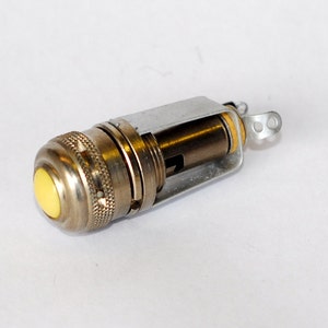 May include: A cylindrical metal electrical component featuring a prominent yellow push button at one end. The body has a textured, knurled grip section and an exposed internal mechanism with a silver-toned metal bracket and two small connection tabs.
