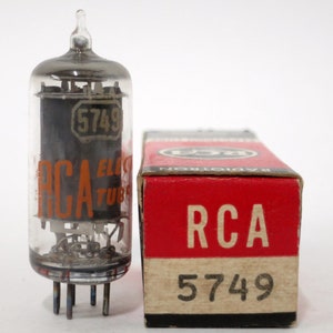 May include: A vintage RCA 5749 vacuum tube with a clear glass enclosure and a black interior. The tube has a series of pins at the base and is accompanied by its original red and beige cardboard box, which also features the RCA logo and the number 5749.