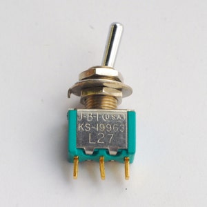 May include: A small, green toggle switch with a silver lever. The switch has the text "J-B-T (U.S.A.) KS-19963 L27" printed on it.