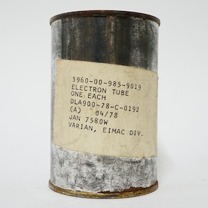 May include: A cylindrical metal container with a label that reads "ELECTRON TUBE." The label includes text such as "ONE EACH" and other technical details. The container has a weathered appearance with signs of age.