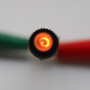 May include: Close-up of a black electrical connector with a glowing orange light inside. The connector is surrounded by green and red electrical wires.