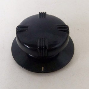 May include: A circular black control knob featuring a raised top section with four evenly spaced sets of vertical ridges along its edge. The base of the knob is a flat, wider circular disc with a single small white indicator line etched into the rim.