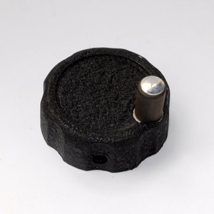 May include: Black plastic knob with a textured surface and a silver metal pin. The knob has a small hole in the center.