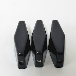 May include: Three black, diamond-shaped objects with a glossy finish are arranged against a white background. Each object has a hole at the bottom and a small, circular indentation. The objects appear to be made of a hard material.