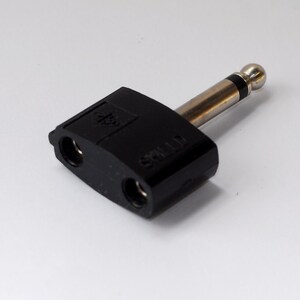 May include: A black audio adapter with two female jacks and a male 1/4 inch (0.6 cm) plug. The adapter has the word "SHIELD" printed on the side. The plug is silver with a black band near the tip.