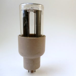 May include: A vintage vacuum tube featuring a clear glass cylindrical top section revealing internal dark metal plates and a sturdy, matte beige ceramic base with small metal pins protruding from the bottom for electrical connection.