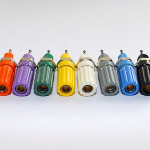 May include: A collection of eight colorful bottle stoppers. Each stopper has a cylindrical body and a small, rounded top. The colors include orange, purple, green, yellow, white, gray, blue, and black. The stoppers are arranged in a row against a white background.