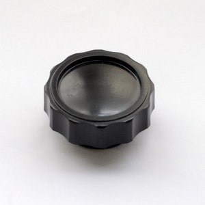 May include: A black, circular object with a ridged edge and a recessed center. The object appears to be made of metal and is set against a white background. The design suggests a functional component, possibly a knob or cap.