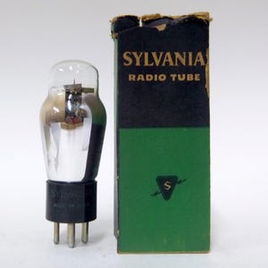 May include: A vintage Sylvania radio tube with a clear glass bulb and black base, next to its original box. The box is black and green with the text "SYLVANIA RADIO TUBE". The tube has metal pins.