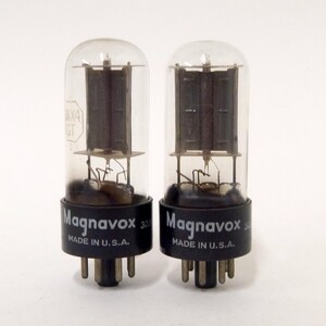 May include: Two vintage Magnavox vacuum tubes. Each tube has a clear glass enclosure with internal components and a black base. The text "Magnavox" and "MADE IN U.S.A." are printed on the base. The tubes are standing upright.