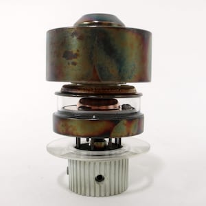 May include: A vintage electronic vacuum tube with a copper and silver colored metal casing. The tube has a clear glass section in the middle and a white plastic base with a threaded ring.