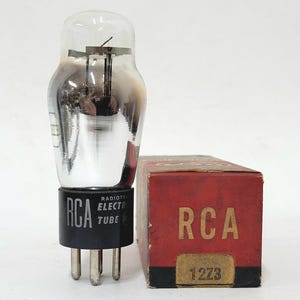 May include: A vintage RCA vacuum tube with a clear glass bulb and black base, standing next to a red box with "RCA" and "12Z3" printed on it. The tube has a metallic internal structure.