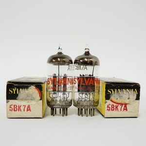 May include: Two vintage Sylvania 5BK7A vacuum tubes with glass enclosures and metal bases. The tubes are next to their original boxes, which have black and yellow labels with red text.