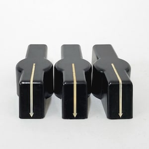 May include: Three black control knobs with a gold vertical line and arrow. The knobs are rectangular with rounded edges and are set against a white background. These knobs are likely for audio equipment or similar electronics.