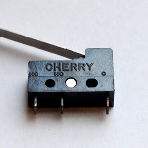 May include: A black Cherry microswitch with a silver lever arm. The switch has three pins and the word "CHERRY" is printed on the front. The letters "NO" are printed on the front of the switch.