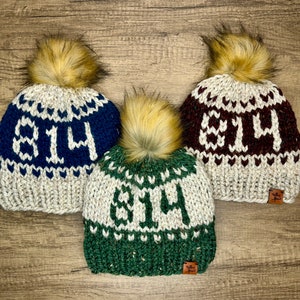 May include: Three knitted hats with pom-poms in different colors. Each hat has the number "814" knitted into the design. The hats are beige with blue, green, and burgundy accents.