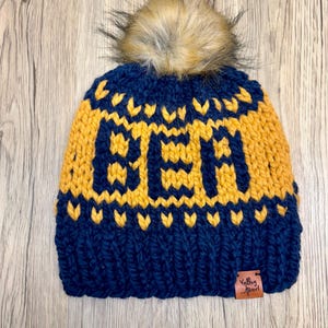May include: A navy blue knit beanie with a yellow knit pattern spelling "YETI" and a brown faux fur pom pom on top. The beanie has a brown leather tag with the text "Valley Pearl" on it.