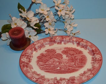 May include: A pink and white oval serving platter with a floral border and a scene of a farmhouse in a wooded area.