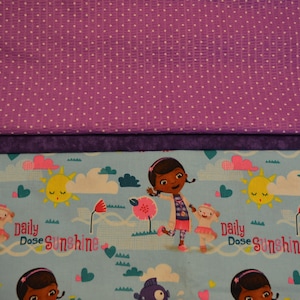 May include: Two pieces of fabric. The top piece is purple with white polka dots. The bottom piece is blue with a repeating pattern of a cartoon girl, a sun, clouds, and the text "daily dose sunshine".