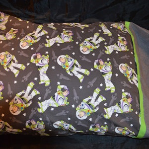 Puede incluir: Una funda de almohada gris con un borde verde presenta un patr&oacute;n repetido de Buzz Lightyear de la franquicia Toy Story. El patr&oacute;n est&aacute; sobre un fondo gris oscuro con estrellas blancas.