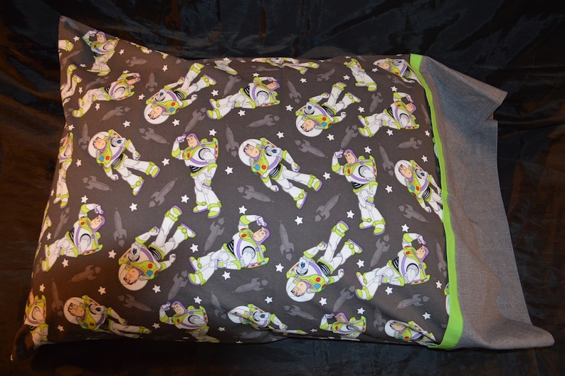 Puede incluir: Una funda de almohada gris con un borde verde presenta un patr&oacute;n repetido de Buzz Lightyear de la franquicia Toy Story. El patr&oacute;n est&aacute; sobre un fondo gris oscuro con estrellas blancas.