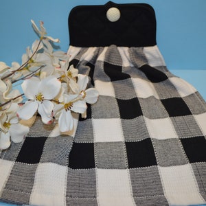 May include: Black and white checkered kitchen towel with a black hanging loop and a white button. The towel is folded and displayed on a blue background.