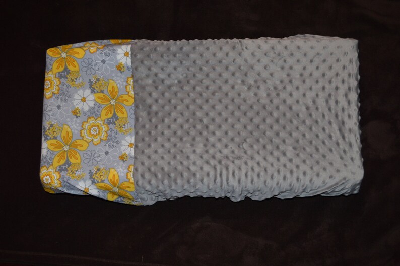 May include: A grey and white baby blanket with a yellow floral pattern on one side. The blanket is made of a soft, textured fabric.