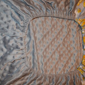 May include: A grey fitted cot sheet with a white and grey quilted pattern. The sheet has a floral print border with yellow and white flowers.