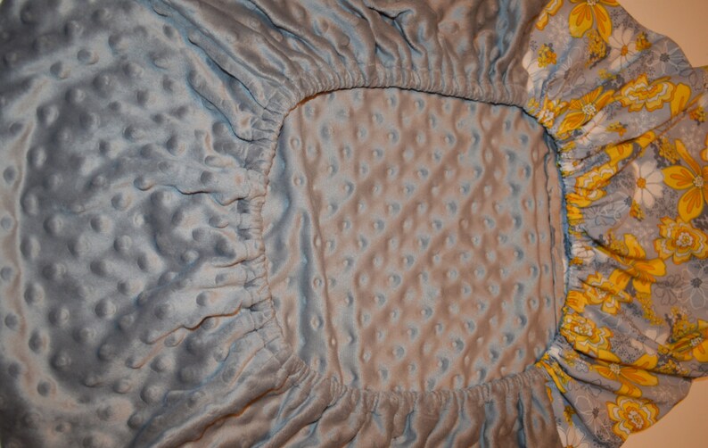 May include: A grey fitted cot sheet with a white and grey quilted pattern. The sheet has a floral print border with yellow and white flowers.