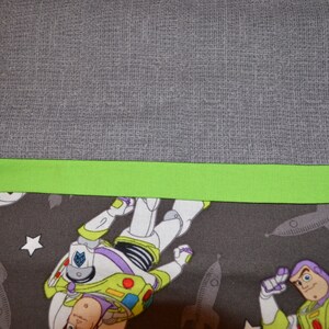 Puede incluir: Primer plano de una tela gris con un ribete verde. La tela tiene un patr&oacute;n de estrellas y naves espaciales con una imagen de dibujos animados de Buzz Lightyear.